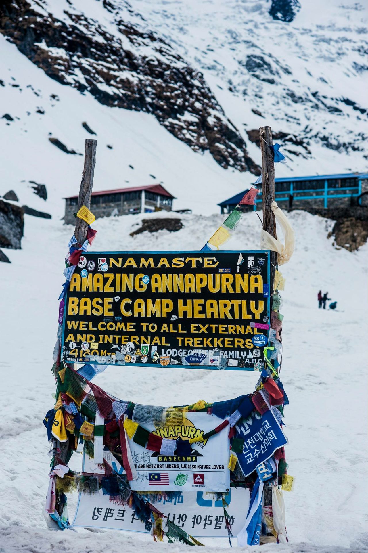 A trailside sign welcoming trekkers to Annapurna Base Camp, snow peaks behind
