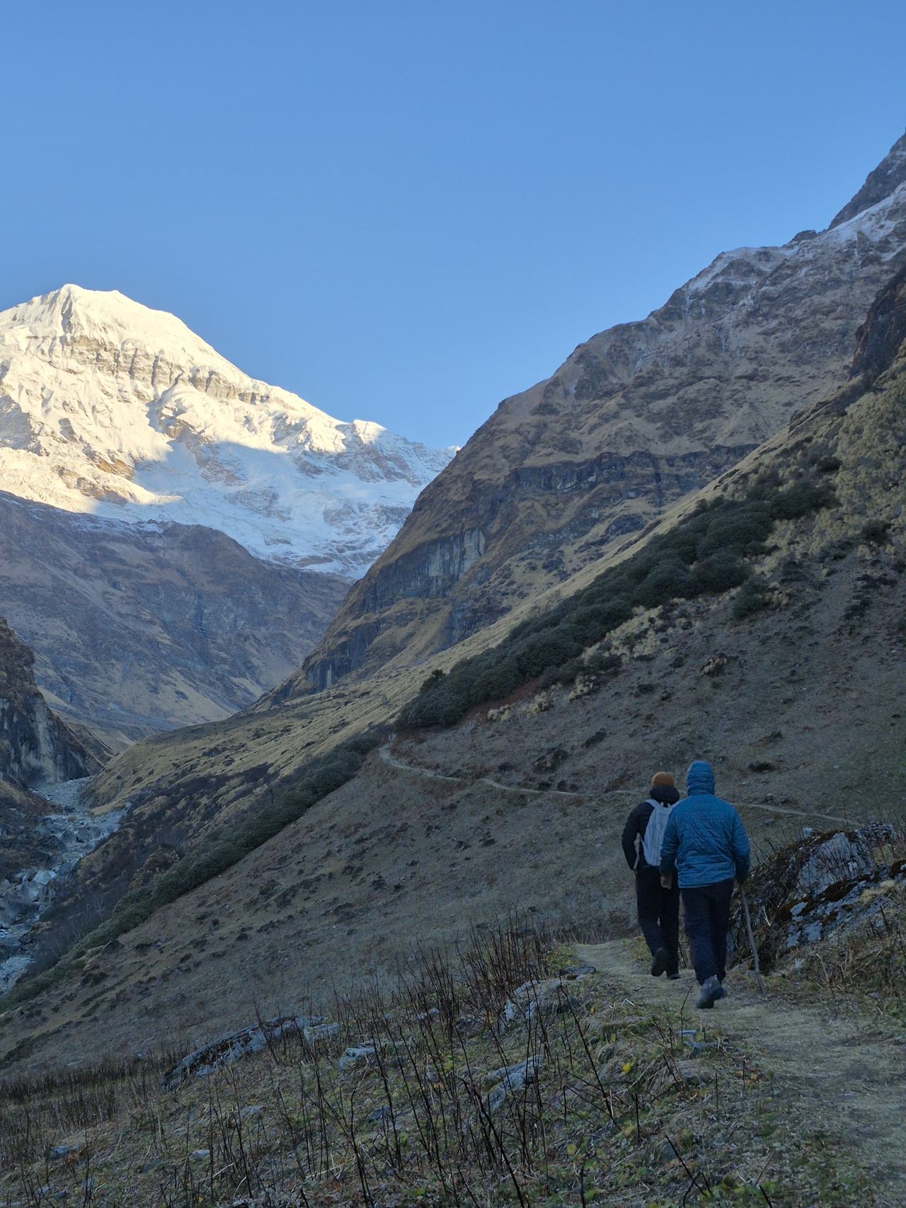 Two trekkers on a mountain trail below a sunlit snow peak