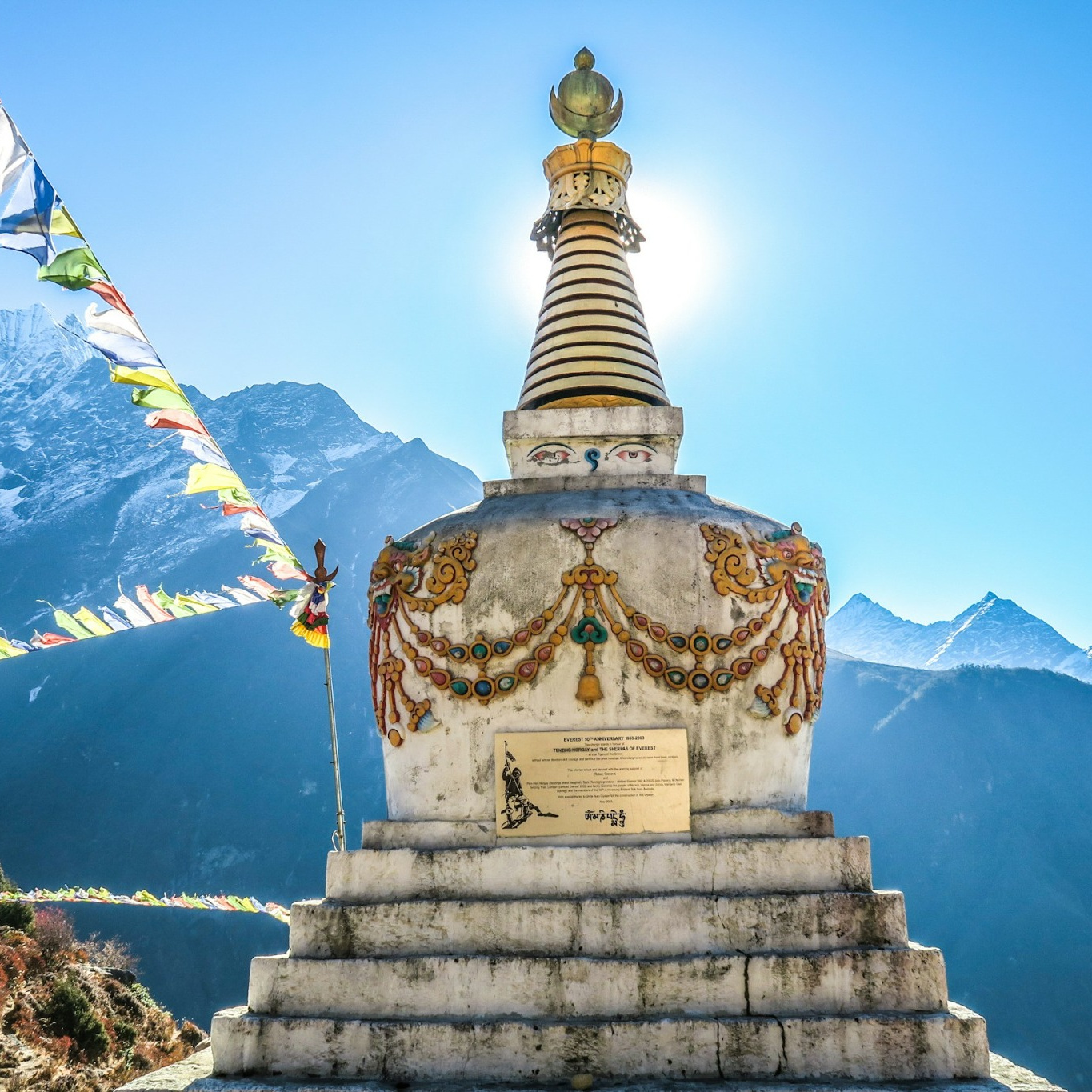 A Buddhist stupa's spire backlit by the sun, prayer flags strung beside it